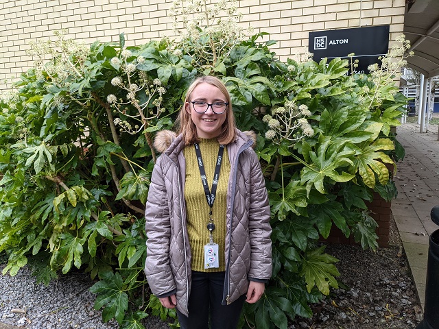 Caroline is smiling and stood in front of a bush